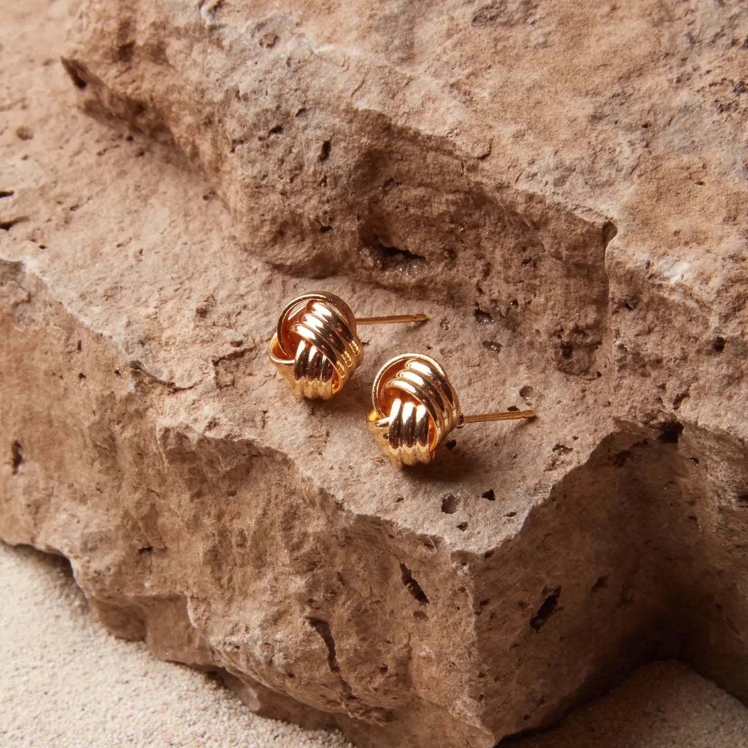 small gold knot stud earrings sitting on a rock