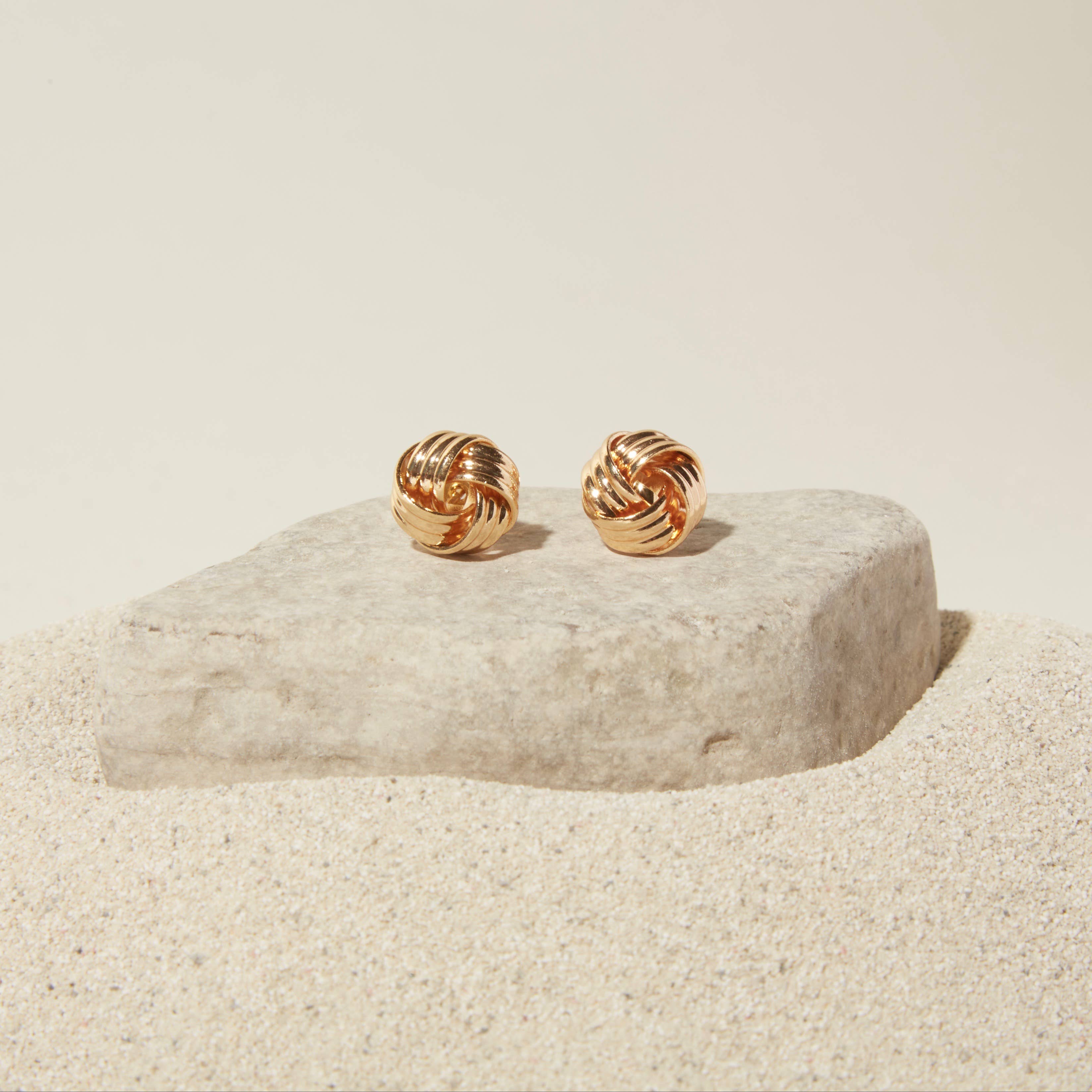 small gold knot stud earrings sitting on a rock in the sand